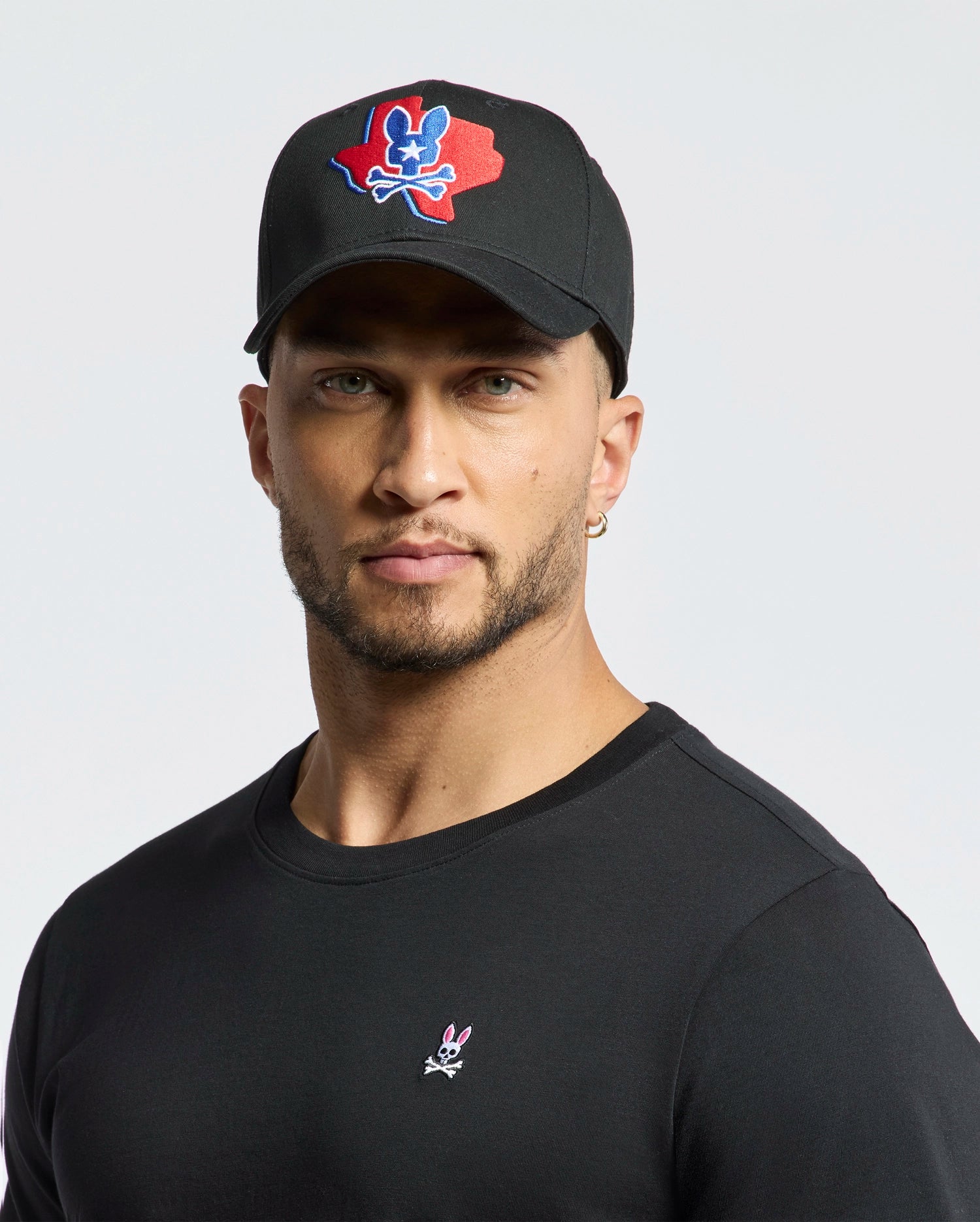 A man dons Psycho Bunny's MENS TEXAS BASEBALL CAP - MO1500014 with a colorful logo and a black cotton shirt embellished with a small design, set against a plain white background.