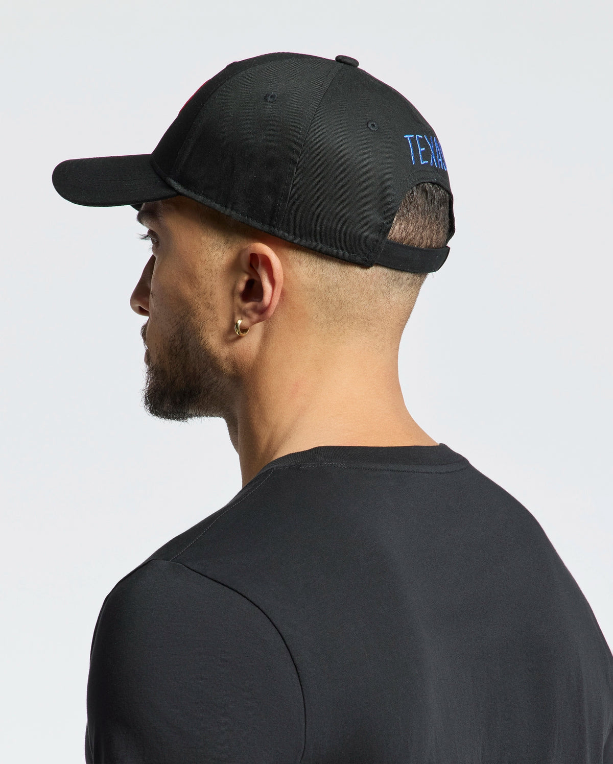A bearded man is seen from the side and back, wearing a black cotton T-shirt and the MENS TEXAS BASEBALL CAP - MO1500014 by Psycho Bunny. The cap features blue woven text on the back. His head slightly turns left against a plain white background.