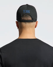 A person with short hair wears a Psycho Bunny "MENS TEXAS BASEBALL CAP - MO1500014," beautifully embroidered in blue on the back. They sport a black cotton t-shirt, an earring in the left ear, and stand against a light gray backdrop.