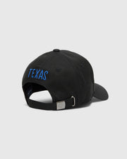 The Psycho Bunny MENS TEXAS BASEBALL CAP - MO1500014 features "TEXAS" embroidered in blue on a black cotton cap. It has a woven adjustable strap with a sleek metal buckle against a plain, light gray background.