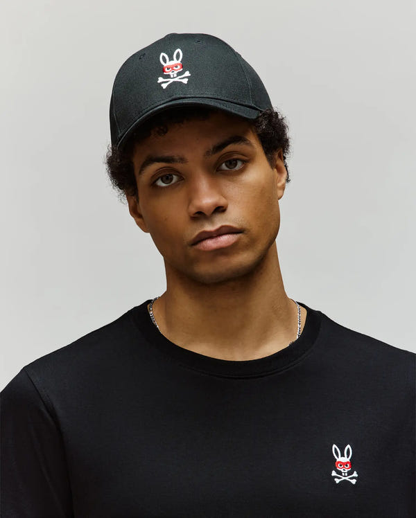 A person in a Psycho Bunny MENS MISCHIEF BASEBALL CAP (MO1500015) with embroidered eyelets and a black t-shirt, both featuring the white Mischief Bunny skull-and-crossbones logo with red eyes, stands against a plain light background.