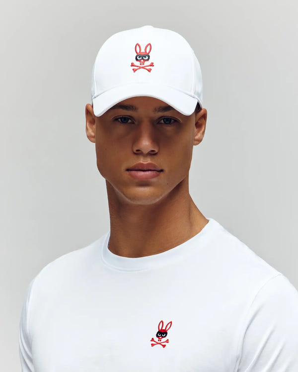 A man wearing a white shirt and the Psycho Bunny MENS MISCHIEF BASEBALL CAP (MO1500015), both featuring the red and black Mischief Bunny with crossbones logo, stands against a light gray background.