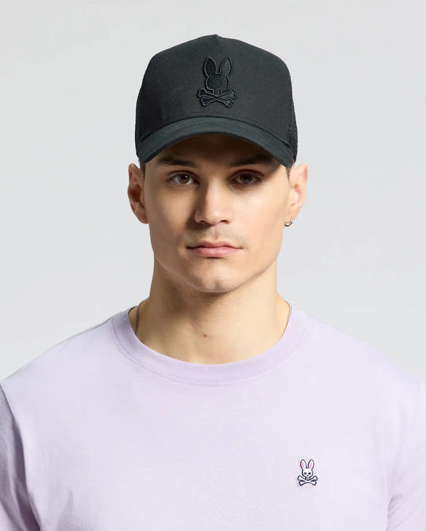 A man wears the Psycho Bunny Men's Wilder Trucker Cap in black (MO1500026) with a bunny skull logo and a pastel purple shirt, featuring a small bunny logo. The adjustable strap ensures a perfect fit as he gazes forward against a neutral backdrop, blending rugged style with playful elements.