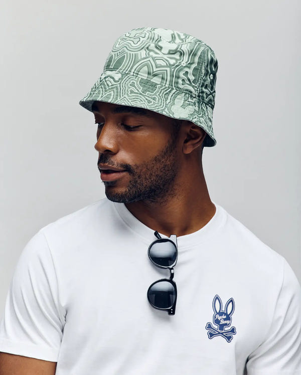 A man wears the Psycho Bunny MENS CHUCK REVERSIBLE BUCKET HAT (MO1500028) in light green pattern and a white t-shirt with a blue Big Bunny logo. Sunglasses hang from his neckline as he looks left against a plain background.
