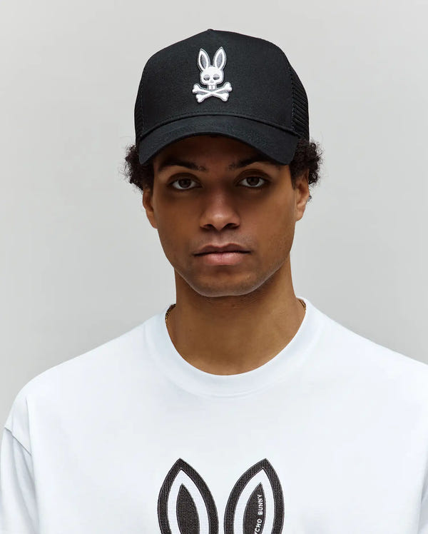 A person wears the Psycho Bunny MENS RYDER TRUCKER CAP - MO1500030, a black mesh snapback with a white bunny skull and crossbones logo, paired with a white shirt featuring a black bunny graphic, against a plain light background.