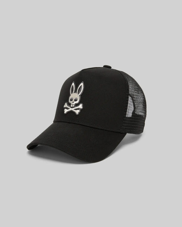 The Psycho Bunny MENS RYDER TRUCKER CAP (MO1500030) is a black cotton twill cap with mesh back, featuring a white embroidered rabbit head and crossed bones on the front, and an adjustable snapback, shown on a light gray background.