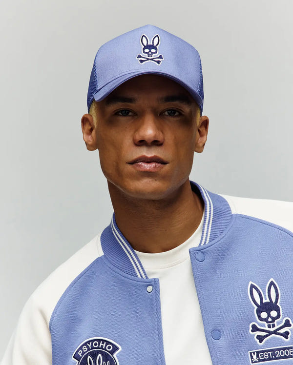 A person poses against a neutral background wearing the Psycho Bunny MENS RYDER TRUCKER CAP (MO1500030) in light blue, featuring a cartoon bunny with crossbones logo and an adjustable cotton twill snapback for the perfect fit.