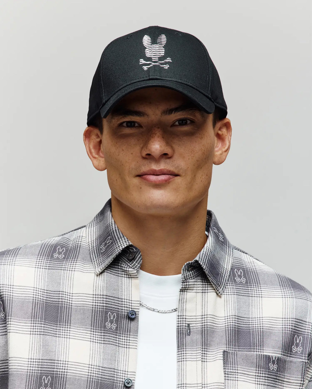 A young man wears the Psycho Bunny MENS GRANT BASEBALL CAP (MO1500037), a black cotton twill cap with a white bunny skull-and-crossbones logo and adjustable backstrap, paired with a plaid shirt featuring small Bunny logos, against a plain background.