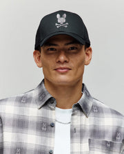 A young man wears the Psycho Bunny MENS GRANT BASEBALL CAP (MO1500037), a black cotton twill cap with a white bunny skull-and-crossbones logo and adjustable backstrap, paired with a plaid shirt featuring small Bunny logos, against a plain background.