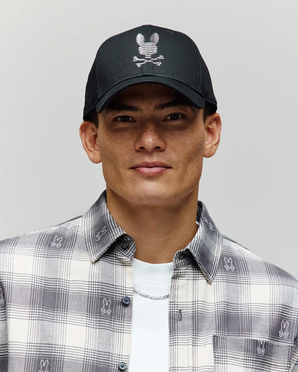 A young man wears the Psycho Bunny MENS GRANT BASEBALL CAP (MO1500037), a black cotton twill cap with a white bunny skull-and-crossbones logo and adjustable backstrap, paired with a plaid shirt featuring small Bunny logos, against a plain background.