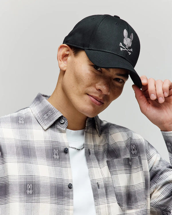 A man in a plaid shirt tips the Psycho Bunny MENS GRANT BASEBALL CAP - MO1500037, featuring a black cotton twill design with a white skull and bunny logo, while smiling against a plain light background.