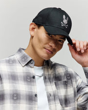 A man in a plaid shirt tips the Psycho Bunny MENS GRANT BASEBALL CAP - MO1500037, featuring a black cotton twill design with a white skull and bunny logo, while smiling against a plain light background.