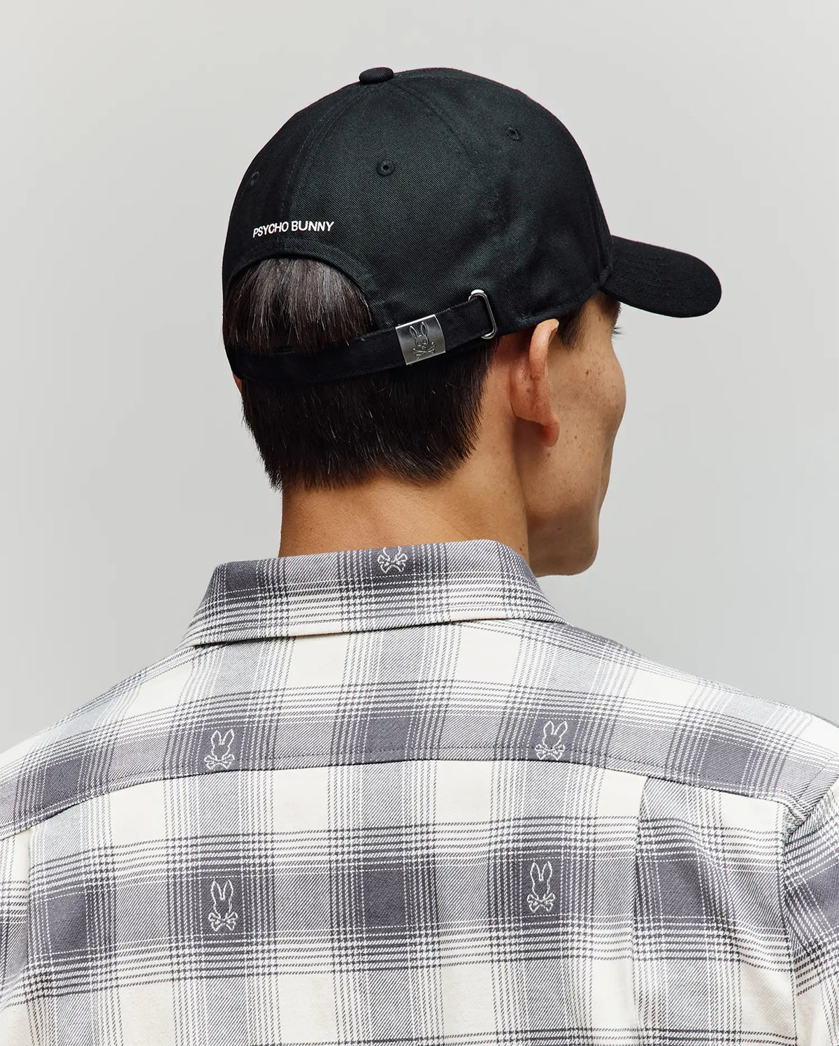 A person wearing a Psycho Bunny MENS GRANT BASEBALL CAP - MO1500037 in black, featuring cotton twill and an adjustable backstrap, along with a plaid shirt with small bunny logos, photographed from behind against a neutral background.
