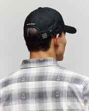 A person wearing a Psycho Bunny MENS GRANT BASEBALL CAP - MO1500037 in black, featuring cotton twill and an adjustable backstrap, along with a plaid shirt with small bunny logos, photographed from behind against a neutral background.