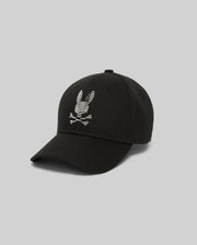 The Psycho Bunny MENS GRANT BASEBALL CAP (MO1500037) is a black cotton twill cap with an embroidered white skull and crossbones with rabbit ears on the front and an adjustable backstrap, shown on a plain gray background.