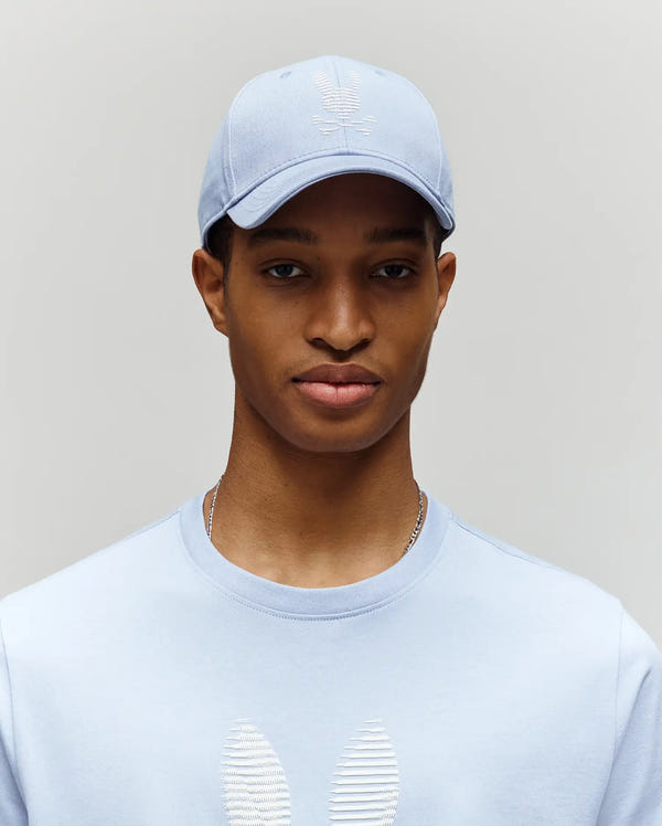 A person wearing the Psycho Bunny MENS GRANT BASEBALL CAP - MO1500﻿037 in light blue, paired with a matching embroidered sweatshirt, stands against a plain light gray background.