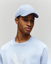 A young man wears the Psycho Bunny MENS GRANT BASEBALL CAP - MO1500﻿037 in light blue with an adjustable backstrap, paired with a matching sweater featuring a white textured bunny logo, standing calmly against a plain background.