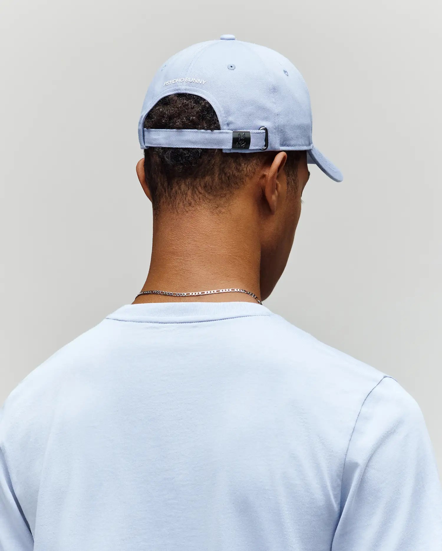 A person is seen from behind wearing a light blue T-shirt and the Psycho Bunny MENS GRANT BASEBALL CAP - MO1500﻿037, a cotton cap with an adjustable backstrap and white text on the back, set against a plain background.