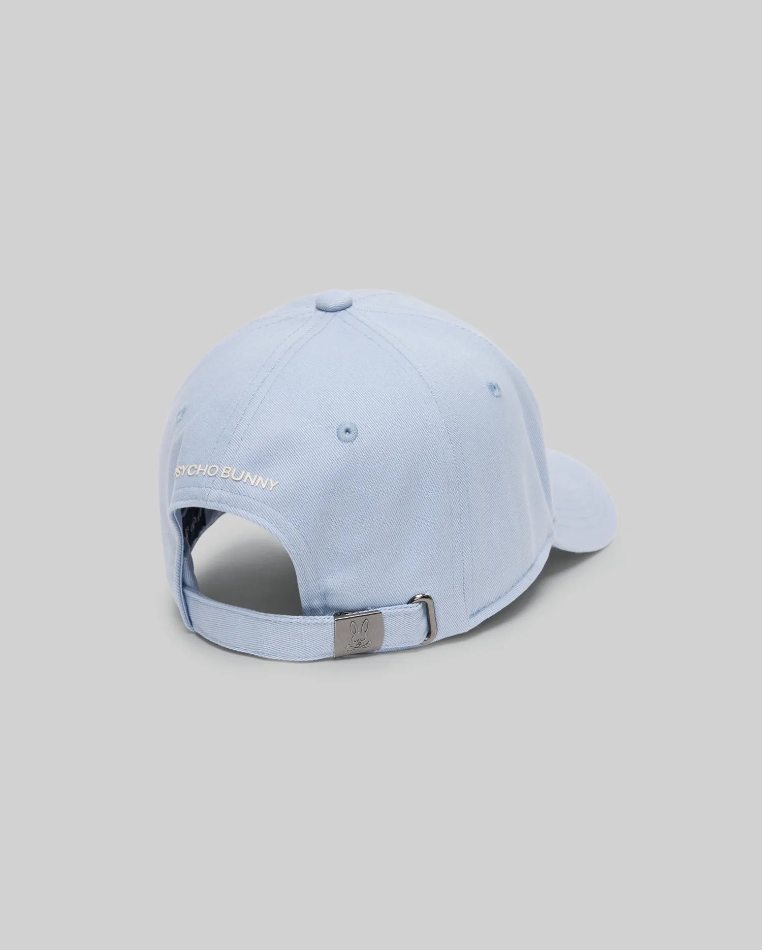 The Psycho Bunny MENS GRANT BASEBALL CAP - MO1500﻿037 is a light blue cotton cap shown from the back, with an adjustable metal clasp backstrap and "PSYCHO BUNNY" embroidered above the opening. The background is plain light gray.