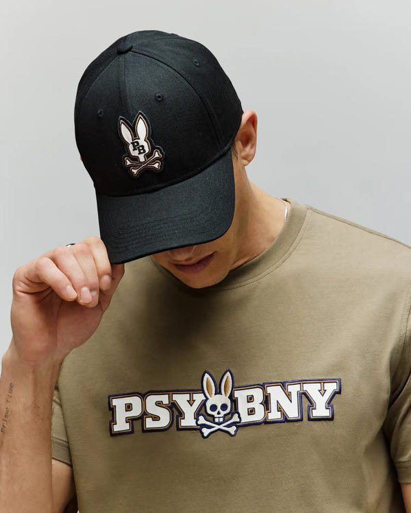 A person wearing the Psycho Bunny MENS WADE BASEBALL CAP (MO1500039), a black cotton twill cap with a bunny skull patch and adjustable backstrap, tilts their head down while holding the brim.