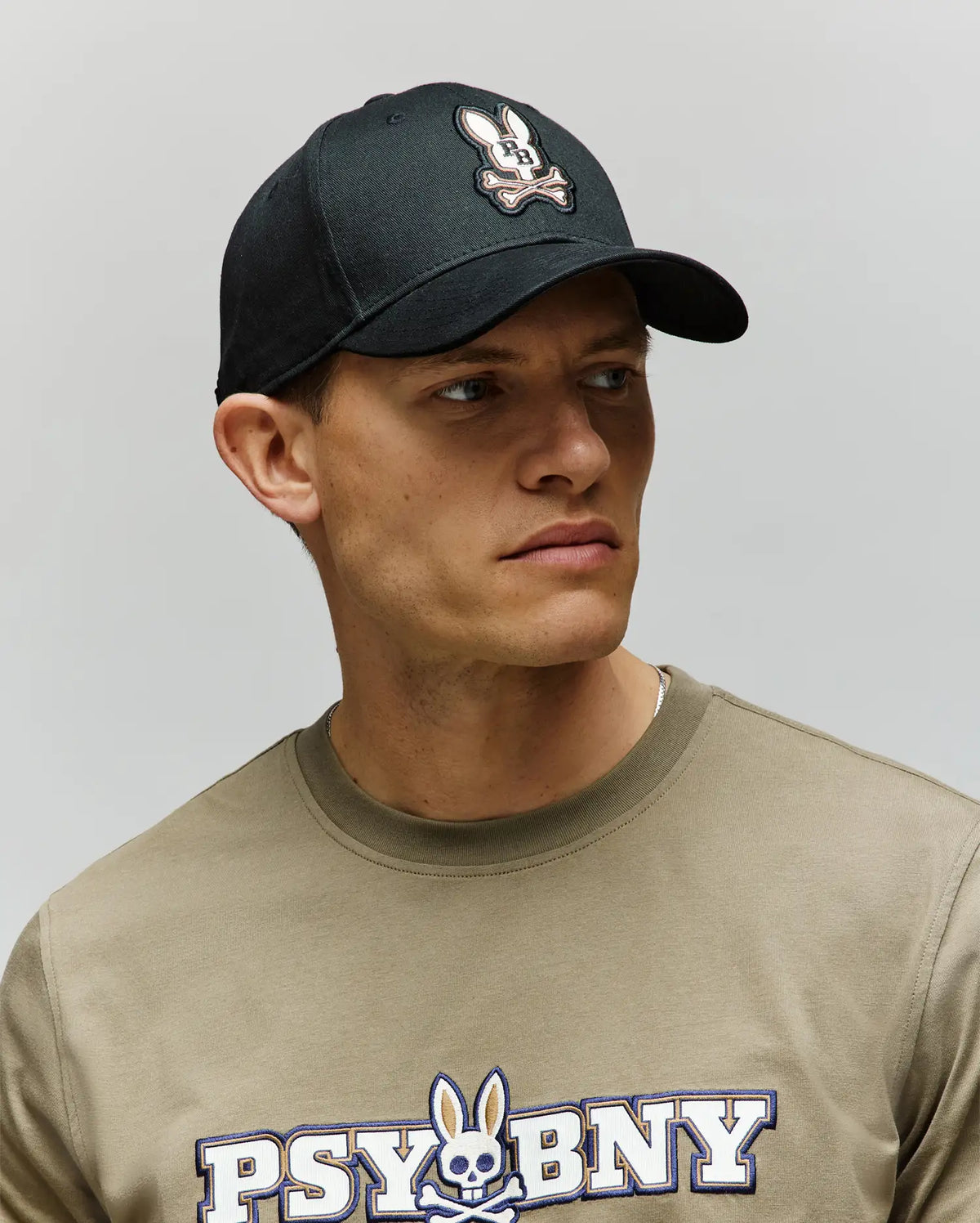 A man wears a Psycho Bunny MENS WADE BASEBALL CAP - MO1500039 in black cotton twill with an embroidered Bunny patch, paired with a matching khaki "PSYCHO BUNNY" T-shirt, looking off to the side against a plain background.