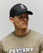 A man wears a Psycho Bunny MENS WADE BASEBALL CAP - MO1500039 in black cotton twill with an embroidered Bunny patch, paired with a matching khaki "PSYCHO BUNNY" T-shirt, looking off to the side against a plain background.