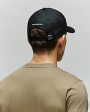 A person seen from the back wears a beige t-shirt and a Psycho Bunny MENS WADE BASEBALL CAP (MO1500039) in black with "PSYCHO BUNNY" embroidered above the adjustable backstrap against a plain light background.