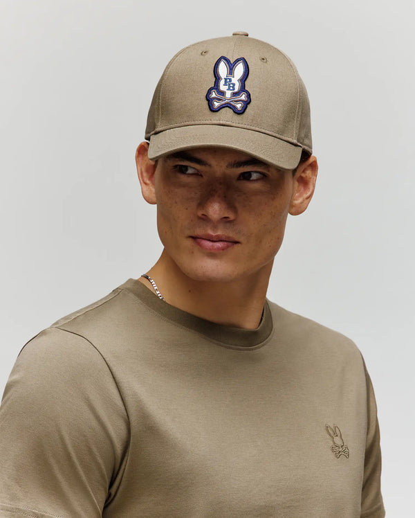 A young man looks to the side against a plain background, wearing the Psycho Bunny MENS WADE BASEBALL CAP - MO1500039 in taupe cotton twill with an embroidered Bunny patch and adjustable backstrap, paired with a matching taupe tee.