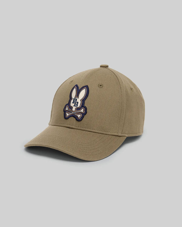 The Psycho Bunny MENS WADE BASEBALL CAP (MO1500039) is a khaki cotton twill cap with an embroidered Bunny patch, crossed bones, "BB" on the front, and an adjustable backstrap, shown on a light gray background.