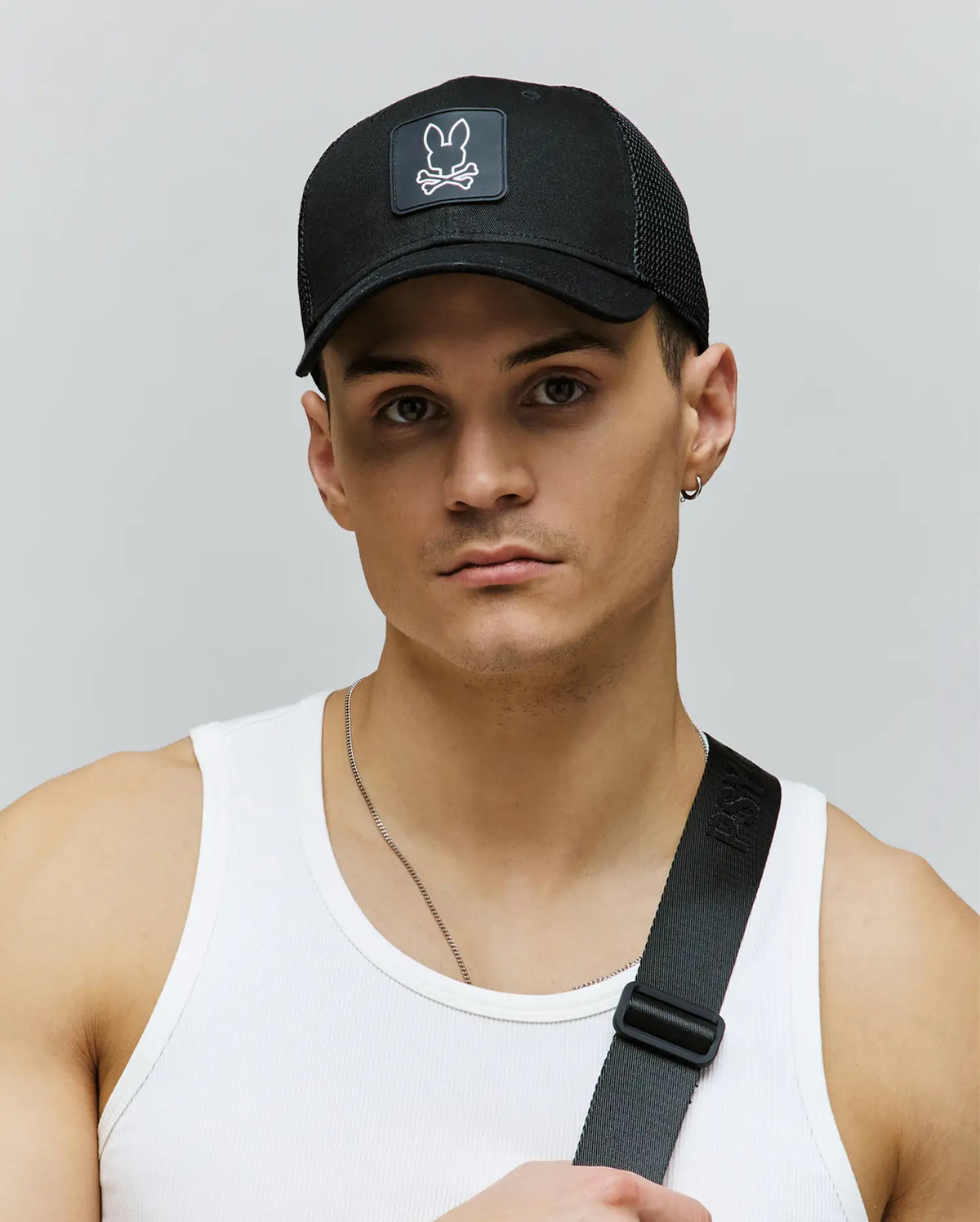 A young man wears the Psycho Bunny MENS DARIO TRUCKER CAP (MO1500044), a black breathable cap with an adjustable snap-back and a white skull and bunny logo, paired with a white tank top and black shoulder strap against a plain background.