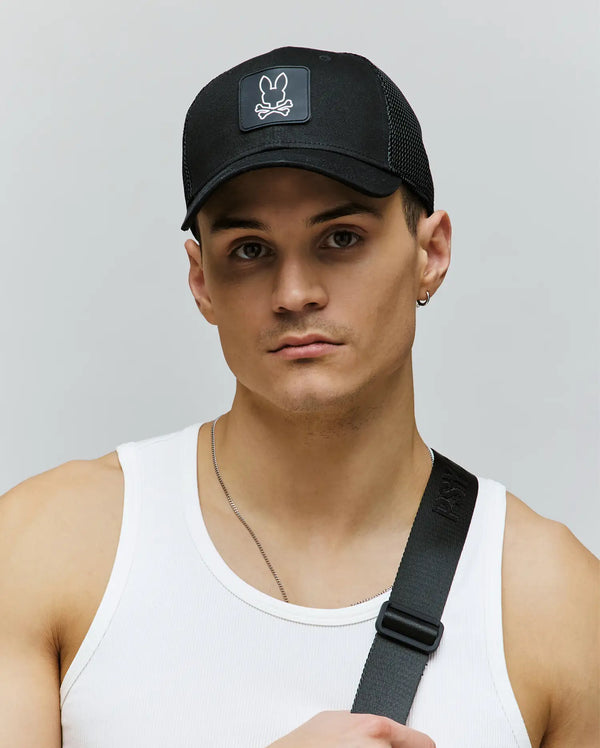 A young man wears the Psycho Bunny MENS DARIO TRUCKER CAP (MO1500044), a black breathable cap with an adjustable snap-back and a white skull and bunny logo, paired with a white tank top and black shoulder strap against a plain background.
