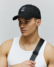 A young man wears the Psycho Bunny MENS DARIO TRUCKER CAP (MO1500044), a breathable black snap-back with a white bunny logo, paired with a white tank top, thin necklace, and black crossbody strap against a plain background.