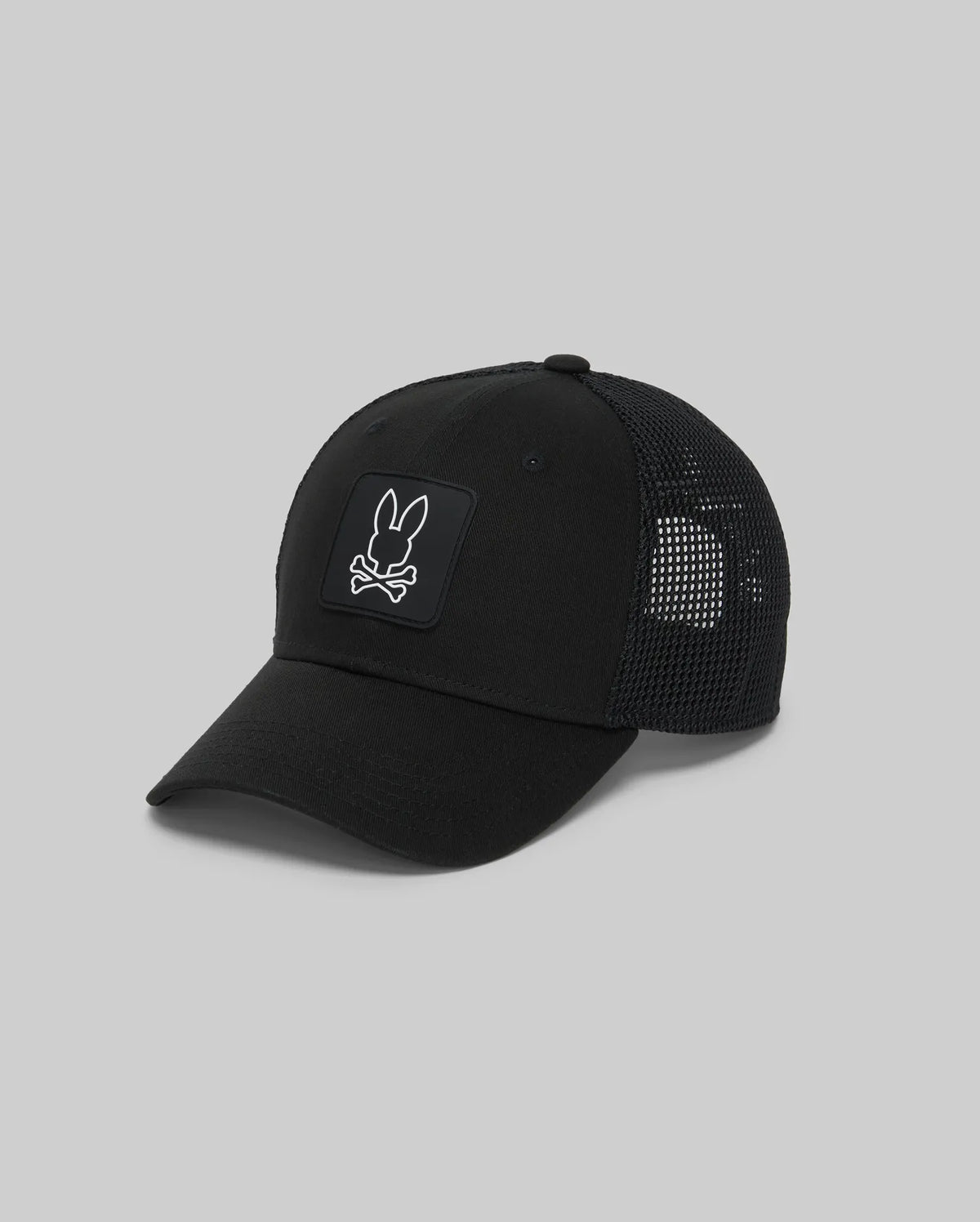 The Psycho Bunny MENS DARIO TRUCKER CAP (MO1500044) is a black cotton twill cap with a mesh back, adjustable snap-back, and features a front patch of a rabbit head above crossed bones on a plain gray background.
