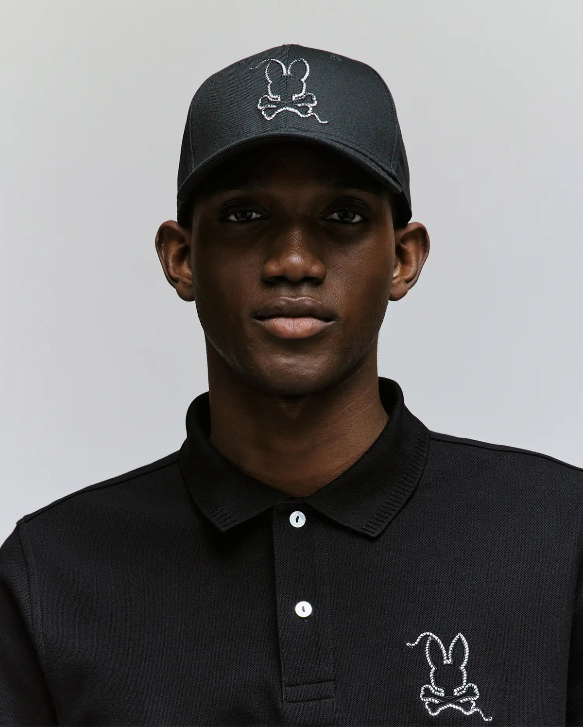 Wearing the Psycho Bunny MENS CYRUS BASEBALL CAP (MO1500045) and a matching black polo, each adorned with a white-outlined bunny logo, a man poses against a light backdrop, bringing subtle coastal charm to his laid-back style.