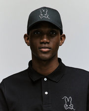 Wearing the Psycho Bunny MENS CYRUS BASEBALL CAP (MO1500045) and a matching black polo, each adorned with a white-outlined bunny logo, a man poses against a light backdrop, bringing subtle coastal charm to his laid-back style.