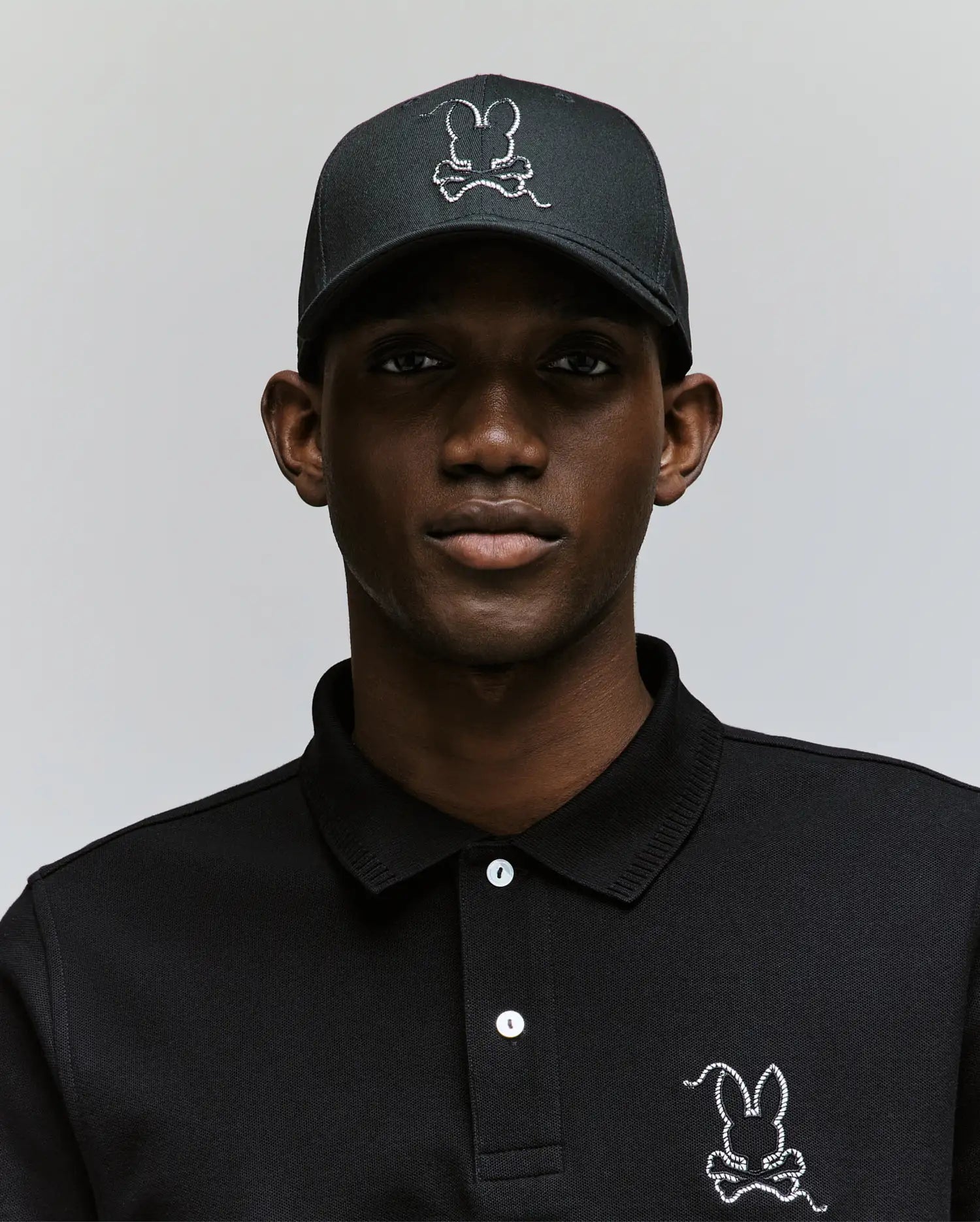 Wearing the Psycho Bunny MENS CYRUS BASEBALL CAP (MO1500045) and a matching black polo, each adorned with a white-outlined bunny logo, a man poses against a light backdrop, bringing subtle coastal charm to his laid-back style.