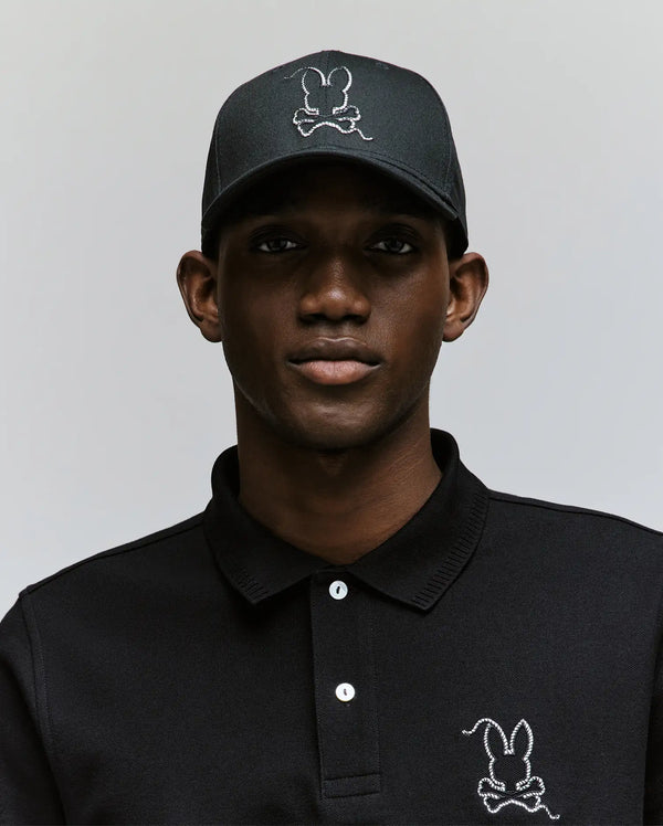 Wearing the Psycho Bunny MENS CYRUS BASEBALL CAP (MO1500045) and a matching black polo, each adorned with a white-outlined bunny logo, a man poses against a light backdrop, bringing subtle coastal charm to his laid-back style.