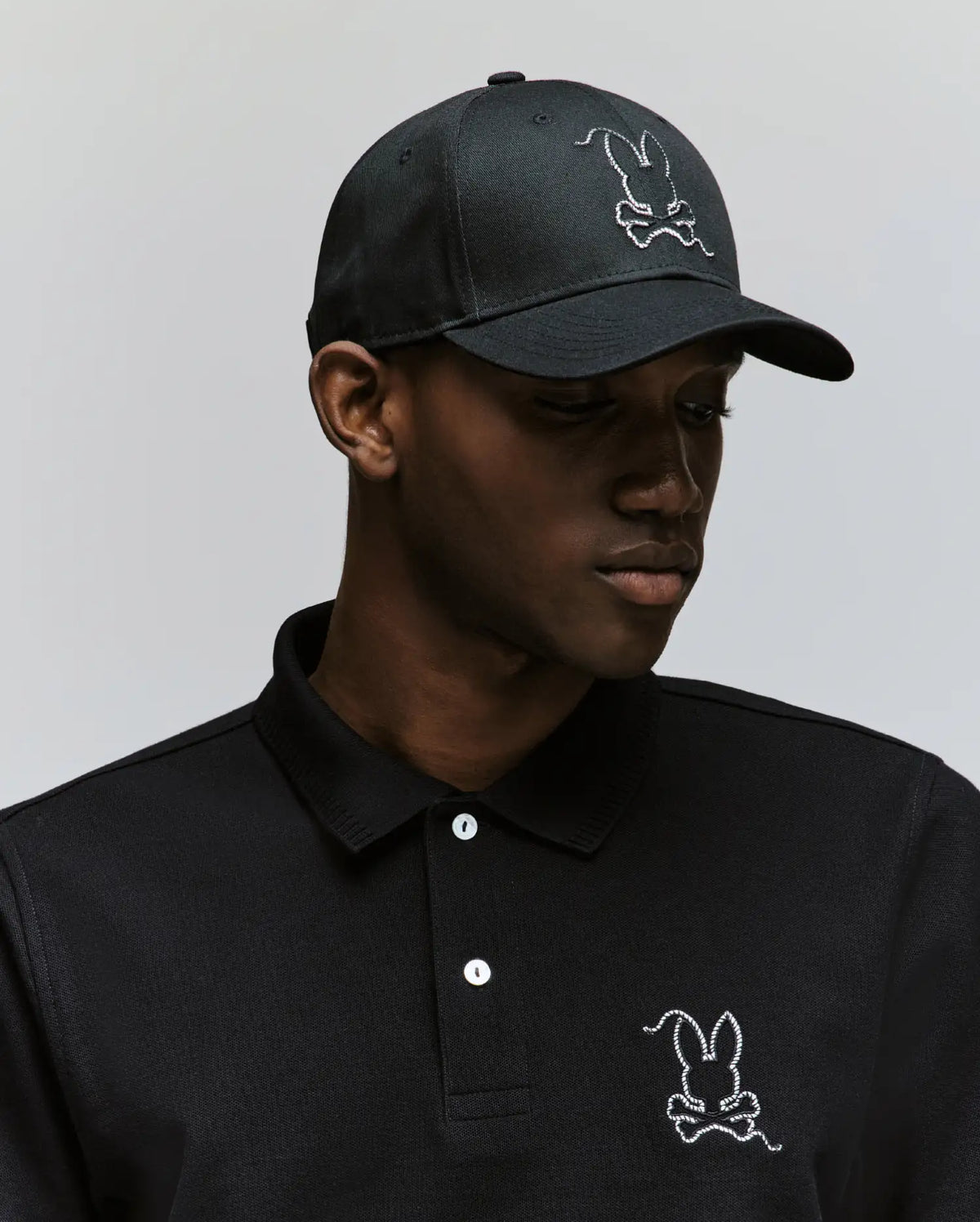 A man wearing a black Psycho Bunny polo and the MENS CYRUS BASEBALL CAP (MO1500045), both featuring a white cartoon bunny and crossbones logo, looks slightly downward against a plain background, adding coastal charm to his look.