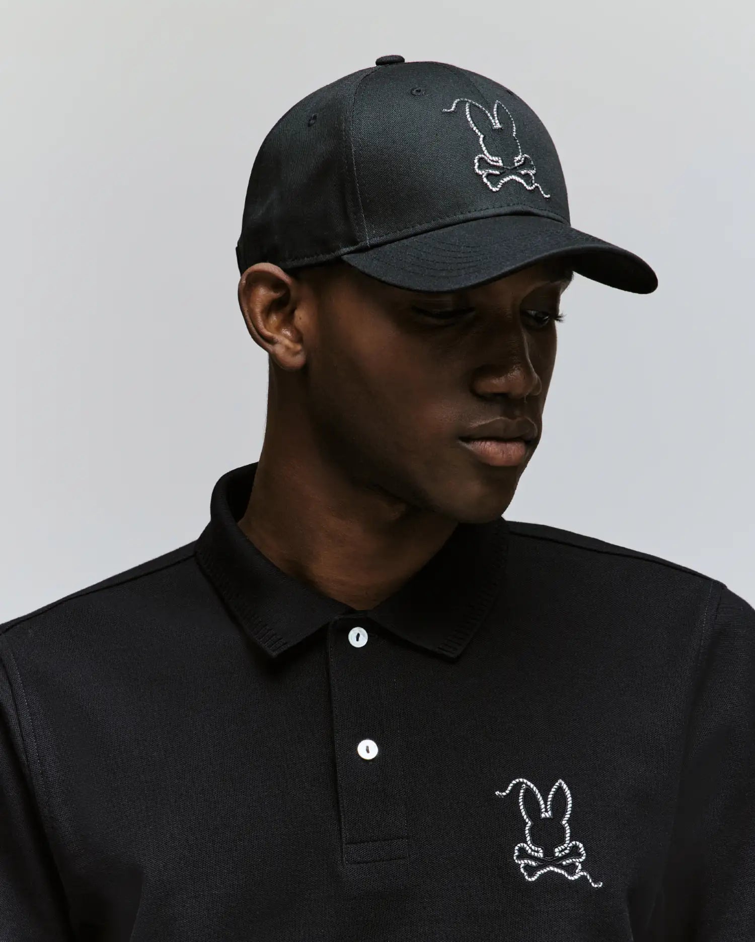 A man wearing a black Psycho Bunny polo and the MENS CYRUS BASEBALL CAP (MO1500045), both featuring a white cartoon bunny and crossbones logo, looks slightly downward against a plain background, adding coastal charm to his look.