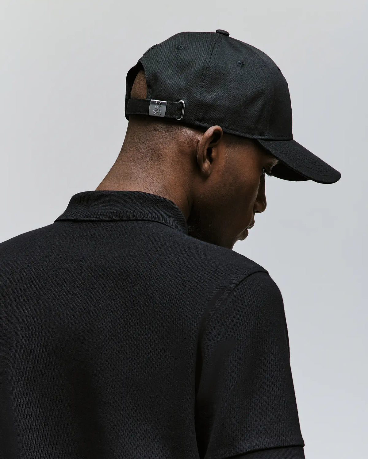 A man is shown from behind wearing the Psycho Bunny MENS CYRUS BASEBALL CAP (MO1500045) with an adjustable metal clasp and a black collared shirt, set against a plain light gray background, embodying subtle coastal charm.