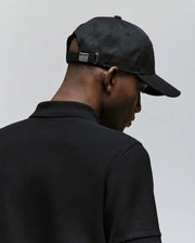 A man is shown from behind wearing the Psycho Bunny MENS CYRUS BASEBALL CAP (MO1500045) with an adjustable metal clasp and a black collared shirt, set against a plain light gray background, embodying subtle coastal charm.