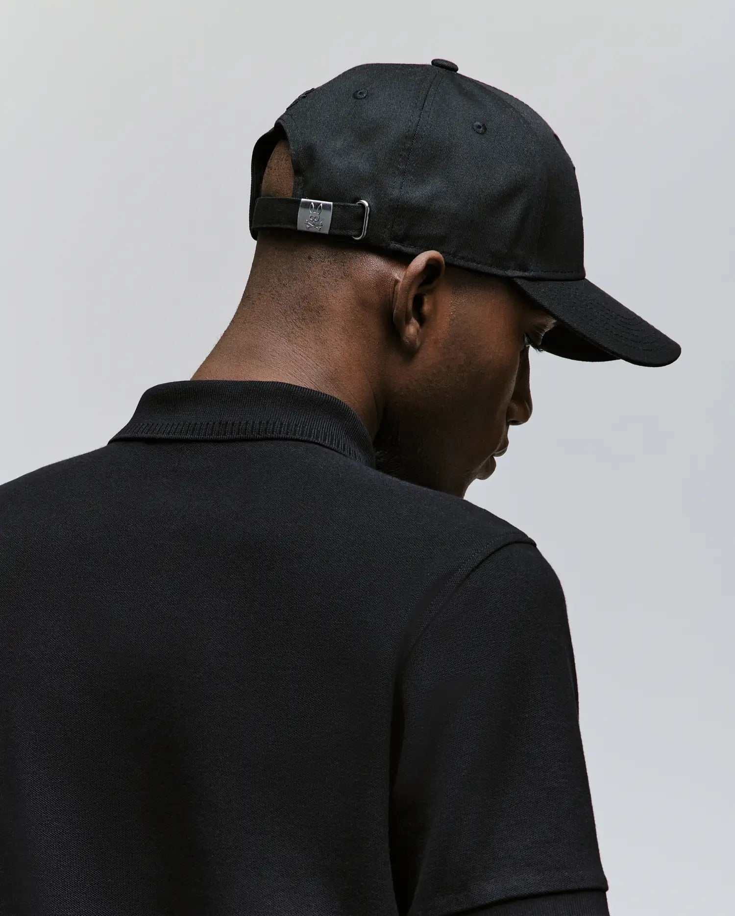 A man is shown from behind wearing the Psycho Bunny MENS CYRUS BASEBALL CAP (MO1500045) with an adjustable metal clasp and a black collared shirt, set against a plain light gray background, embodying subtle coastal charm.