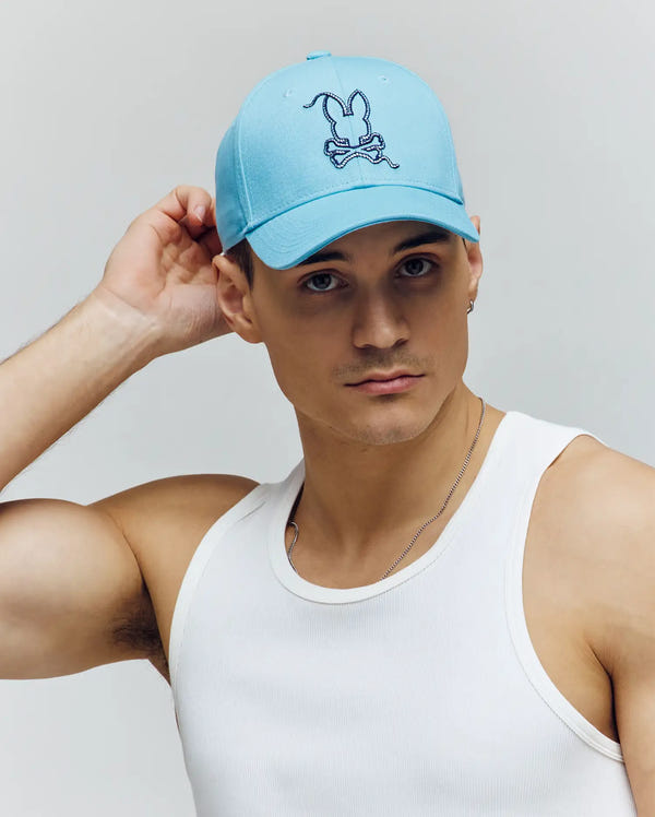 A man in a white tank top poses against a plain light background, wearing the Psycho Bunny MENS CYRUS BASEBALL CAP (MO1500045), featuring the brand's signature bunny and crossbones design, with one hand resting on his cap.