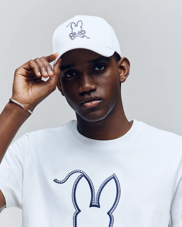 A person in a white Psycho Bunny MENS CYRUS BASEBALL CAP (MO1500045) featuring the bunny and crossbones logo, along with a matching white t-shirt, stands against a plain grey background.