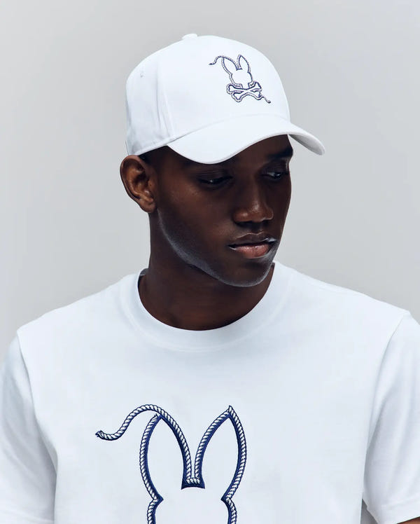 A man in the Psycho Bunny MENS CYRUS BASEBALL CAP (MO1500045) and a matching tee, both with a navy bunny and crossbones outline, adds subtle coastal charm as he looks down to the right against a light gray background.