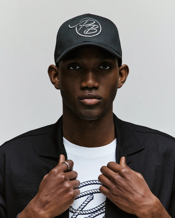 A man wears the Psycho Bunny MENS CYRUS PB BASEBALL CAP - MO1500047 in black and a matching jacket over a white Cyrus tee with a rope-style logo, looking ahead while holding his jacket against a neutral background.