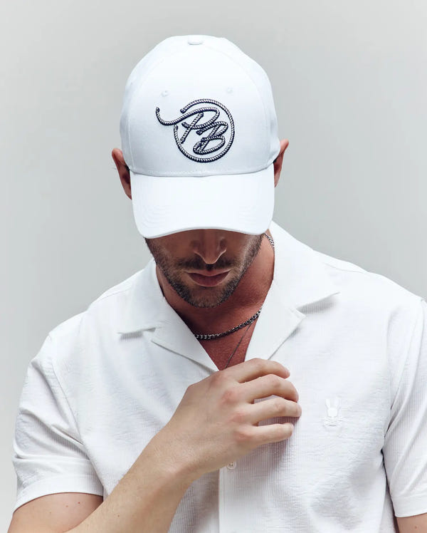 A man wearing the Psycho Bunny MENS CYRUS PB BASEBALL CAP (MO1500047), a white short-sleeve shirt, and a silver chain necklace looks down with one hand near his collar against a plain, light background.