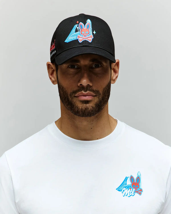 A bearded man wears a Psycho Bunny MENS MONTREAL BASEBALL CAP (MO1500048) and a white 100% cotton shirt, both featuring colorful embroidered Bunny graphics with blue accents, against a plain light grey background.