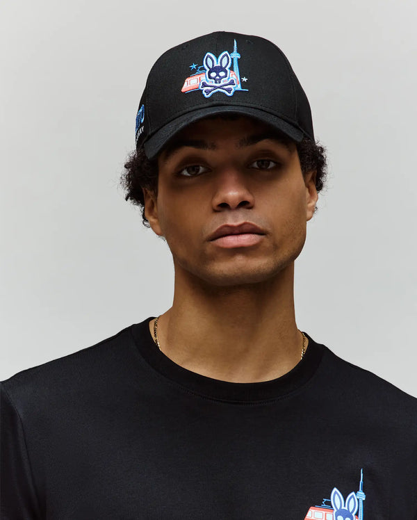 A man wears the Psycho Bunny MENS TORONTO BASEBALL CAP - MO1500049 and a matching tee, both with blue cartoon rabbit and CN Tower graphics, looking at the camera against a plain light background.