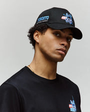 A young man wears the Psycho Bunny MENS TORONTO BASEBALL CAP (MO1500049) with an embroidered Bunny logo and adjustable strap, paired with a black tee featuring a blue and red chest graphic, posing against a plain light background.
